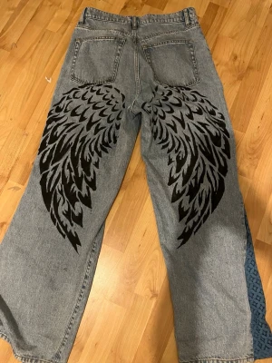 Baggy jeans med vingar och spets - Unika baggy jeans i ljusblå denim med stora svarta vingar målade på baksidan. Utsvängda ben med blå spetsinsats som ger extra edge. Perfekta för dig som vill sticka ut och ha en cool streetstyle-look.