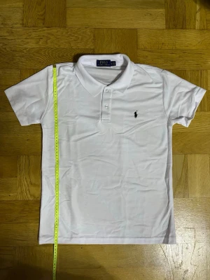 Vit pikétröja från Polo Ralph Lauren - Klassisk vit pikétröja från Polo Ralph Lauren med korta ärmar, krage och knäppning med tre knappar. Tröjan har den ikoniska broderade loggan på bröstet och är gjord i mjukt bomullsmaterial. Perfekt för en clean och stilren look.