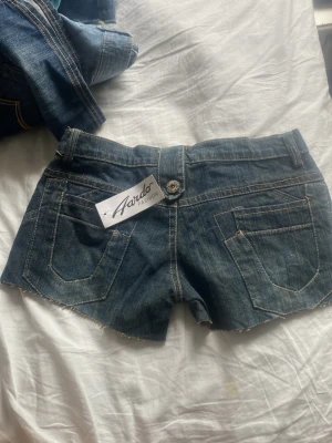 Blå jeansshorts från Aardo Fashion - Säljer ett par blå jeansshorts från Aardo Fashion med råa kanter och snygga detaljer som dekorativa knappar på fickorna. Klassisk femficksmodell med bälteshällor och slitna detaljer för en cool look.