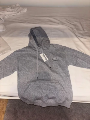 Grå hoodie från CD - Snygg grå hoodie från CD med broderad logga på bröstet. Klassisk passform med huva och dragsnören samt stor magficka framtill. Tillverkad i mjukt bomullsmaterial som känns skönt mot huden. Perfekt för en avslappnad och trendig look.