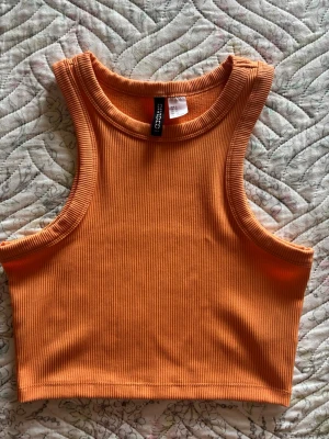 Ribbad orange croppad linnetopp - Säljer en ribbad, croppad linnetopp från Divided i en snygg orange färg. Toppen är ärmlös och har en tight passform med rund halsringning. Perfekt för varma dagar eller som layering-piece.