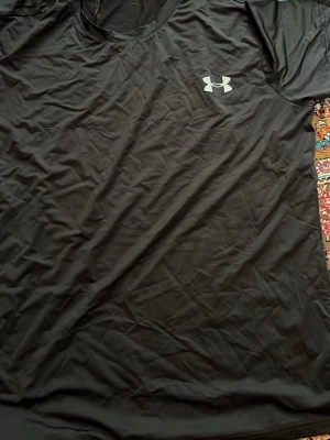 Svart träningströja från Under Armour - Svart kortärmad träningströja från Under Armour med rund hals och logga på bröstet. Tillverkad i ett lätt och snabbtorkande material som passar perfekt för träning och sportaktiviteter.