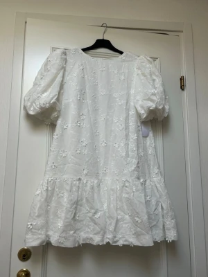 Vit kort klänning med puffärm och spets - Supersöt luftig vit kort klänning med puffärmar och broderade blommor. Klänningen har öppen rygg med knyt och volang nedtill. Endast provad, ej använd, prislapp är kvar
