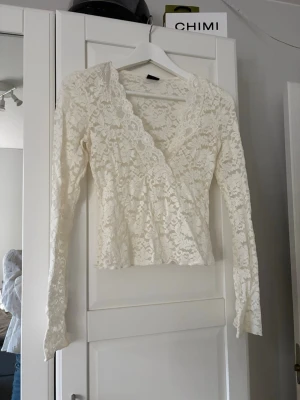 Vit spetsblus från Gina Tricot - Säljer en superfin vit spetsblus från Gina Tricot i omlottmodell. Blusen har långa ärmar, v-ringning och vackra spetsdetaljer över hela plagget. Perfekt att styla med jeans eller kjol för en trendig look.