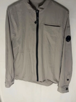 Beige overshirt med dragkedja - Stilren beige overshirt med svart dragkedja framtill och bröstficka. Långärmad modell med knappdetaljer vid ärmslut och en rund svart patch på ena ärmen. Tillverkad i ett lätt och tunt material, perfekt för lager-på-lager.