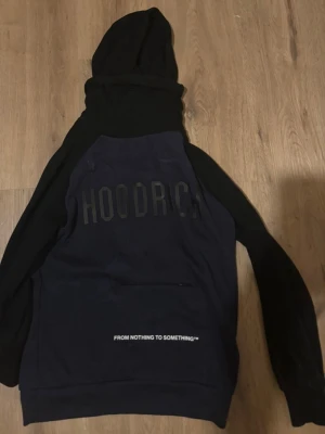 Svart och marinblå HOODRICH hoodie - Snygg hoodie i svart och marinblått från HOODRICH med stor huva och trycket 'HOODRICH' på bröstet. På magfickan står det 'FROM NOTHING TO SOMETHING'. Hoodien har långa ärmar och klassisk känguruficka framtill. Perfekt för en streetwear-look.