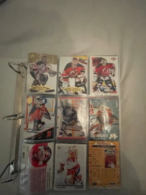 Samling hockeykort i pärm - Säljer en samling hockeykort i pärm med olika målvakter från NHL och internationella lag. Korten har färgstarka motiv och visar spelare i action, flera med klassiska målvaktsutrustningar och lagtröjor. Perfekt för dig som gillar hockey eller vill utöka din kortsamling.