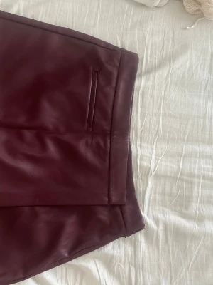 Vinröd kortkjol i skinnimitation - Säljer en vinröd kortkjol från Stradivarius i skinnimitation. Kjolen har en stilren design med fickdetalj framtill och mjukt innerfoder. Perfekt för dig som vill ha en edgy look med en touch av trend.
