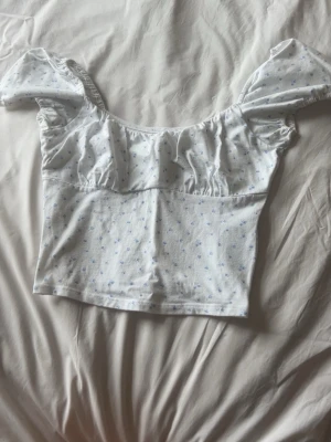 Vit blommig topp med puffärm - Supersöt vit topp med små blå blommor och puffiga korta ärmar. Toppen har en lätt croppad passform och rynkade detaljer vid bysten. Perfekt för dig som gillar romantisk och feminin stil.