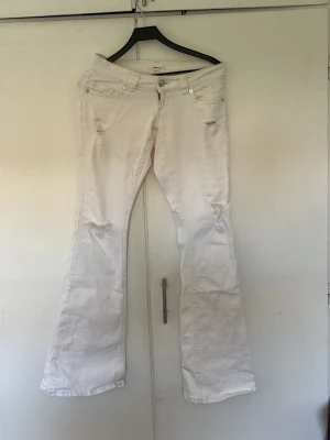 Vita bootcut jeans med slitningar - Säljer ett par vita bootcut jeans med slitna detaljer på låren och knäna. Jeansen har fem fickor, broderade bakfickor och stängs med knapp och dragkedja. Perfekta för dig som gillar en avslappnad och trendig stil.