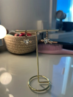 Guldörhängen med hjärtformad sten - Säljer ett par örhängen i guldton med hjärtformad klar sten och små guldpilar på sidorna. Dem har tappat lite färg vid själva krokarna, som synd på bild💕