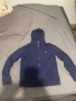 Marinblå hoodie från Polo Ralph Lauren - Säljer en marinblå hoodie från Polo Ralph Lauren med dragkedja, huva och fickor framtill. Klassisk liten logga broderad på bröstet. Skön bomullsmix som passar perfekt till chill eller streetwear. Enkel och stilren design som alltid funkar. Finns en liten fläck som syns på bild vid frågor eller funderingar är det bara att skriva.