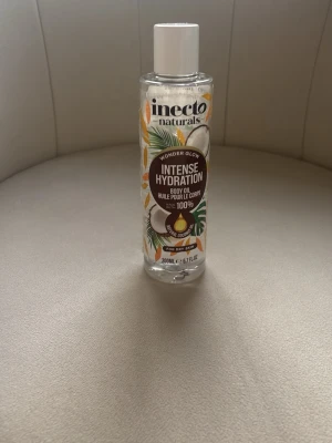 Inecto Intense Hydration Body Oil - Kroppsolja från Inecto med kokosdoft, ger intensiv återfuktning och glow. Kommer i en genomskinlig plastflaska med vitt lock och tropiskt inspirerad etikett. Ändast lite använd som man ser på bild 2  passar torr hud. Oljan är klar och lättflytande.