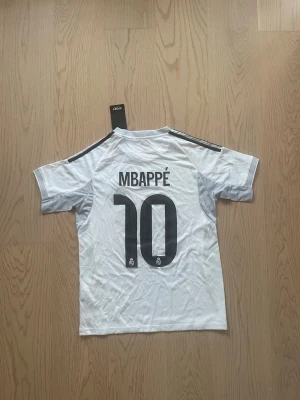 Mbappé Real Madrid hemmaställ - Säljer en vit Real Madrid matchtröja med Mbappé och nummer 10 på ryggen. Tröjan har svarta detaljer på axlarna, klubbmärke och Emirates Fly Better-tryck på bröstet. Korta ärmar och sportig passform, perfekt för fotbollsträning eller match.