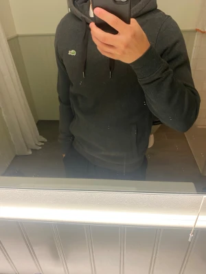 Mörkgrå hoodie från Lacoste - Snygg mörkgrå hoodie från Lacoste med klassisk krokodillogga på bröstet. Hoodien har dragsko i huvan och är tillverkad i mjukt material som känns skönt mot huden. Perfekt för en avslappnad och stilren look. Passar även för dem som har xs