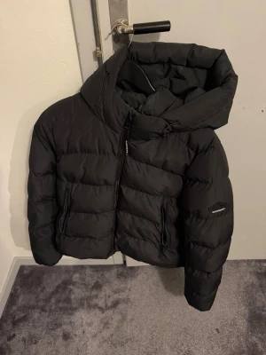 Svart pufferjacka med huva - Säljer en svart pufferjacka med stor huva och dragkedja framtill. Jackan har två snedställda fickor med dragkedja och en patch på ärmen. Perfekt för kalla dagar och riktigt skön att bära. Klassisk boxig passform och quiltad design.