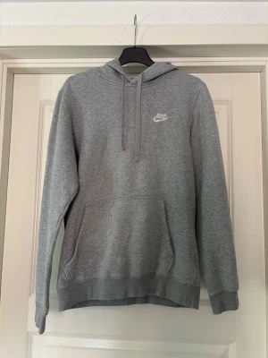 Grå hoodie från Nike med huva - Klassisk grå hoodie från Nike med vit logga på bröstet. Hoodien har huva med dragsko, stor magficka och ribbade muddar vid ärmar och nederkant. Tillverkad i mjukt sweatshirtmaterial som är perfekt för en avslappnad stil.