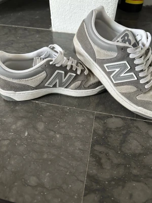 Grå New Balance sneakers  - Snygga grå sneakers från New Balance med klassisk N-logga på sidan. Skorna är låga och har ovandel i mocka och textil, samt vita snören och detaljer. Perfekta för dig som gillar stilrena och bekväma sneakers. 