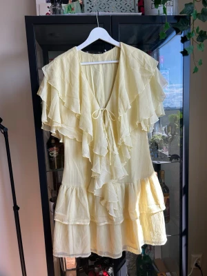 Ljusgul volangklänning med knyt - Supersöt ljusgul kortklänning från Nakd med flera lager volanger och knyt framtill. Klänningen har en luftig passform och är tillverkad i ett lätt, tunt tyg (chiffon) som ger en somrig känsla. Perfekt för dig som gillar romantiska detaljer och en feminin look.