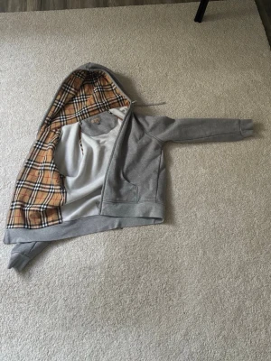 Grå hoodie med rutigt foder i huvan - Snygg grå hoodie med dragkedja och klassisk huva. Insidan av huvan har ett stilrent beige, svart och vitt rutmönster som ger en exklusiv känsla. Hoodien har långa ärmar och ribbade muddar. Perfekt för en chill och trendig look.