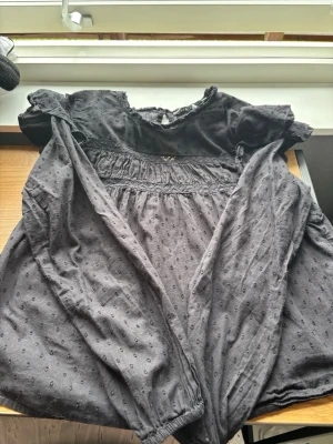 Svart transparent blus med volang - Säljer en svart transparent blus med diskret prickmönster och volangdetaljer vid axlarna. Blusen har rund halsringning, knapp i nacken och långa ärmar med resår vid ärmslut. Perfekt för dig som gillar en luftig och trendig stil.