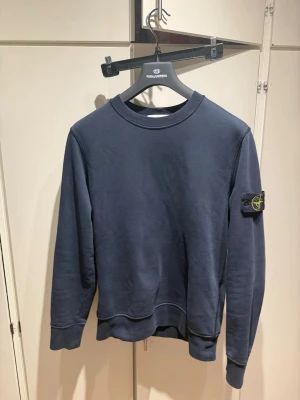 Mörkblå sweatshirt från Stone Island - Mörkblå sweatshirt från Stone Island med klassisk rund halsringning och ribbade muddar. Tröjan har den ikoniska Stone Island-loggan på vänster ärm. Perfekt för en avslappnad och stilren look. Pris är diskuterbart!
