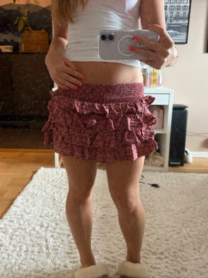 Snygg kortkjol - Supersöt kort kjol i rött med småblommigt mönster och volangdetaljer. Kjolen har bred resår i midjan och flera lager volanger som ger en lekfull look. Perfekt för dig som vill ha en trendig och bekväm kjol till sommaren.
