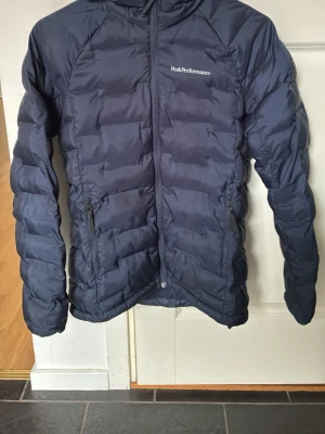 Mörkblå dunjacka från Peak Performance - Snygg mörkblå dunjacka från Peak Performance med quiltad design och dragkedja framtill. Jackan har två sidofickor med dragkedja och en diskret logga på bröstet. Perfekt för kalla dagar och enkel att matcha med olika outfits. Storlek XS. Använd sparsamt 
