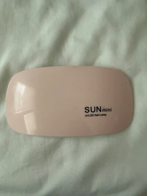 UV LED nagellampa SUN mini - Smidig och kompakt UV LED-lampa för naglar från SUN mini. Lampan är ljusrosa på ovansidan och vit undertill, med fyra LED-lampor för effektiv härdning av gelélack. Perfekt storlek för att ta med sig och enkel att använda hemma.