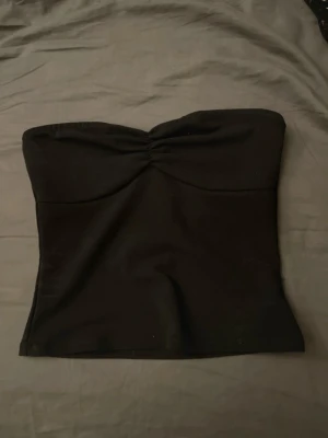 Svart tube topp - Säljer en svart bandeau tubtopp med lätt rynkning framtill. Toppen är axelbandslös och har en enkel, stilren design som passar till många olika looks. 