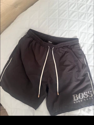 Svarta badbyxor från Hugo Boss - Snygga svarta badbyxor från Hugo Boss med vita dragsnören och logga på vänster ben. Shortsen har elastisk midja och vita detaljer längs sidorna. 