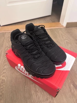 Svarta Nike Air Max Plus sneakers TN  - Nike Air Max Plus sneakers i helsvart med mesh- och syntetmaterial, ikoniska vågformade detaljer och synlig Air Max-dämpning i sulan. Klassisk låg modell med snörning och diskret logga på plösen. Perfekta för en sportig och stilren look.( lite sönder men ett stor håll