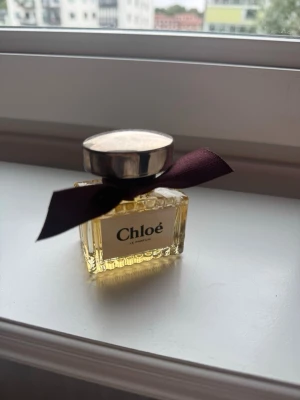 Chloé Le Parfum miniflaska - Söt miniflaska av Chloé Le Parfum med guldfärgad vätska, elegant glasflaska med räfflad design och en stor silverfärgad kork. Flaskan är dekorerad med ett mörklila band knutet runt halsen för en lyxig känsla.