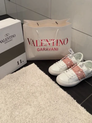 Valentino Rockstud Sneakers - Snygga vita sneakers från Valentino med breda rosa band över vristen dekorerade med ikoniska nitar. Skorna har en chunky sula och är tillverkade i läder med coola detaljer som ger en lyxig vibe. Perfekta för dig som vill sticka ut med stil.