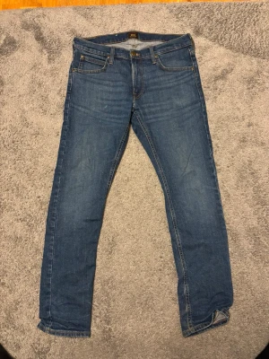 Blå slim fit jeans från Lee - Snygga blå slim fit jeans från Lee med klassisk femficksdesign och läderpatch bak i midjan. Jeansen har normal midja och smala ben, perfekta till sneakers. Tillverkade i mjuk denim med lätt stretch för extra komfort.