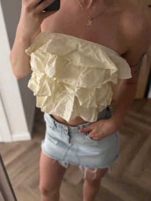 Gul volangig tubtopp - Säljer en ljusgul axelbandslös tubtopp med flera lager volanger. Toppen är croppad och har en luftig och somrig känsla, perfekt till jeanskjol eller shorts. Materialet känns som bomull och toppen är superenkel att styla för en trendig look.
