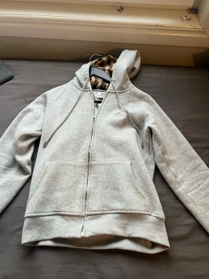 Ljusgrå zip hoodie med huva - Säljer en ljusgrå hoodie med dragkedja och stor ficka framtill. Hoodien har en klassisk huva med dragsko och ribbade muddar vid ärmslut och nederkant. Perfekt för chill dagar eller när du vill ha något bekvämt och stilrent. Med trasig drag kedja