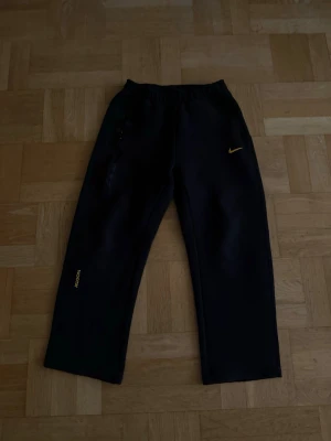 Svarta Nike NOCTA mjukisbyxor - Svarta mjukisbyxor från Nike NOCTA med raka ben och resår i midjan. Byxorna har gula detaljer med NOCTA- och Swoosh-logga samt diskret tryck på benet. Perfekta för en chill och sportig stil.