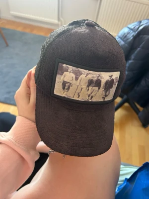 Svart truckerkeps med vintage-tryck - Säljer en svart truckerkeps med mesh baktill och justerbar snapback. Framtill sitter ett vintage-inspirerat tryck med svartvit bild på fem personer. Kepsen har böjd skärm och klassisk form, perfekt för en avslappnad streetwear-look.