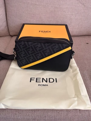 Fendi väska - Snygg axelväska från Fendi i svart med klassiskt FF-mönster och en gul diagonal detalj på framsidan. Väskan har justerbar axelrem och silverfärgade metalldetaljer. Kommer med originalbox och dustbag.