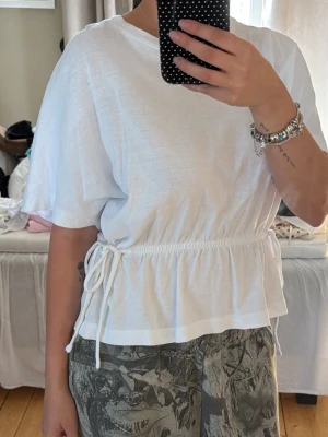 Vit kortärmad topp med knyt - Säljer en vit kortärmad topp med rund hals och knyt i midjan som ger en snygg form. Toppen är i mjuk bomull och har en avslappnad passform. Perfekt att styla med både jeans och kjol.