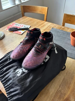 Puma future elite 7 i svart lila färg - Säljer ett par snygga fotbollsskor med högt skaft i svart och lila med röda detaljer. Skorna har snörning och en strumpliknande krage för extra stöd runt ankeln. Ytan är mönstrad för bättre bollkontroll och sulan är anpassad för spel på konstgräs. Påse tillkommer