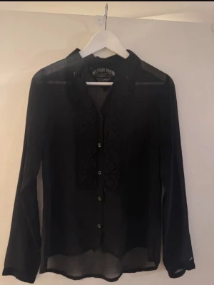Svart blus med volang från Hilfiger Denim - Stilren svart blus från Hilfiger Denim med transparent material och dekorativa volanger längs knappraden framtill. Klassisk krage och långa ärmar ger en elegant look. Perfekt att matcha med jeans eller kjol för en trendig outfit.