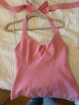Rutig rosa linnetopp med knytband - Supersöt rosa linnetopp med vitt rutmönster och breda knytband i nacken. Toppen har en lätt v-ringad form och är ärmlös, perfekt för varma dagar. Materialet känns som bomull eller polyester och toppen har en figurnära passform.