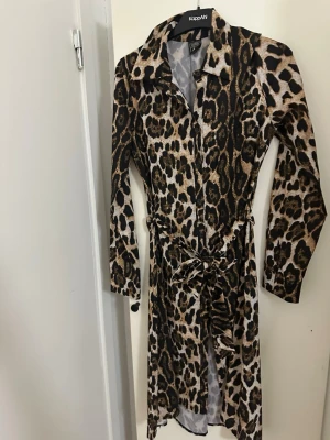 Leopardmönstrad klänning med bälte - Snygg långärmad klänning med leopardmönster och knytbälte i midjan. Klänningen har krage och knappar framtill, vilket ger en cool och trendig look. Perfekt för dig som vill sticka ut med ett djurmönster.