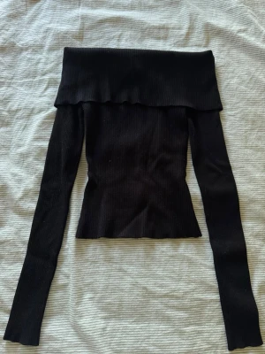 Svart offshoulder topp från H&M - Snygg svart ribbad offshoulder topp från H&M i storlek XS. Toppen har långa ärmar och en liten croppad passform som sitter tight. Lappen har gått av på ena sidan