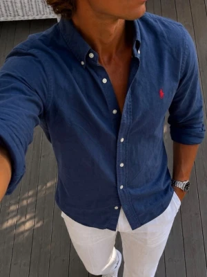Ralph Lauren skjorta med knappar - Snygg mörkblå Ralph Lauren skjorta med klassisk krage och vita knappar. Skjortan har lång ärm och en liten röd broderad logga på bröstet. Perfekt för dig som gillar stilrena och tidlösa plagg.