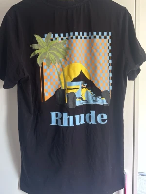Svart Rhude t-shirt med tryck - Cool svart t-shirt från Rhude med färgglatt tryck på ryggen som visar en palm, solnedgång och en bil framför rutigt mönster. På bröstet finns en mindre logga med palm och bokstaven R. Perfekt för dig som gillar streetwear och unika prints.