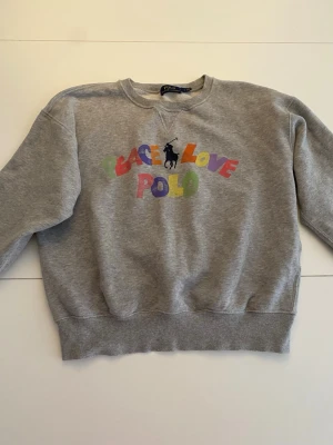 Grå sweatshirt från Polo Ralph Lauren - Grå sweatshirt med färgglad text 'Peace Love Polo' Tröjan har rund halsringning, ribbade muddar och avslappnad passform. Perfekt för en chill och trendig look.storlek S men passar xs också,aldrig använd!!