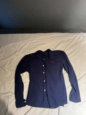 Marinblå skjorta från Polo Ralph Lauren - Klassisk marinblå skjorta från Polo Ralph Lauren med vit knappstängning och röd broderad logga på bröstet. Skjortan har lång ärm och mjukt bomullsmaterial som känns skönt mot huden. Perfekt till jeans eller chinos.